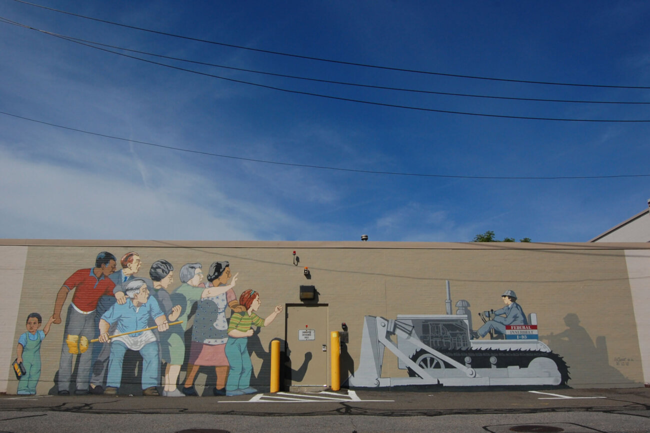 A mural on the side of a cement building showing a diverse group of people squaring off against a man in a bulldozer.