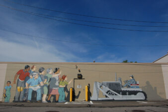 A mural on the side of a cement building showing a diverse group of people squaring off against a man in a bulldozer.