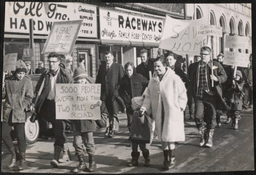 A large group of people of all ages march along a sidewalk holding anti highway signs.