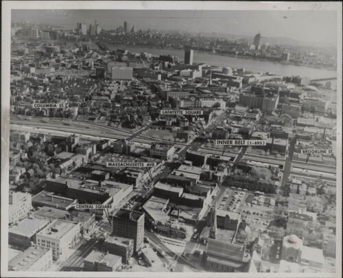 An aerial photo of a city with the route of a highway cutting through the middle. There are several small labels marking different city squares.
