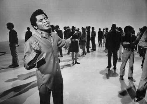 A Black man performing on a set with a loose crowd of people behind him.
