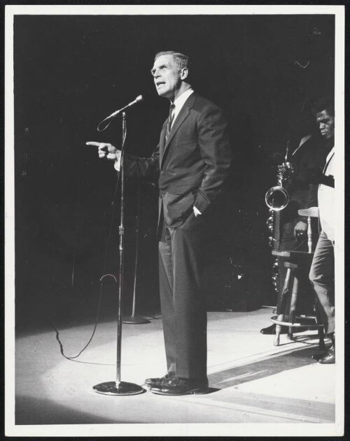 A man in a suit standing at a microphone with his finger pointing at the audience. There's a man with a saxophone behind him in a white suit.
