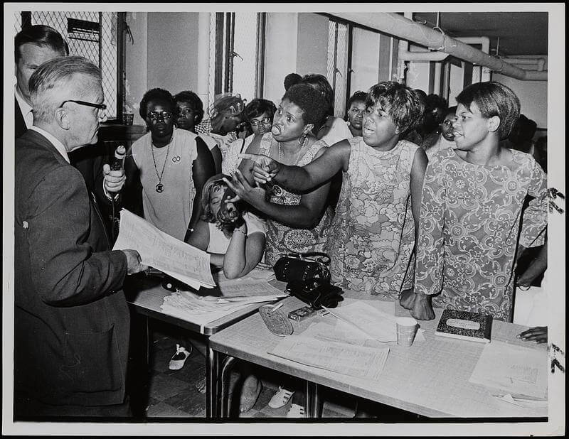 A group of Black women arguing with a white man behind a desk.