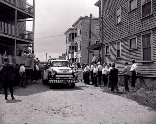 An emergency vehicle going down a narrow street with men in uniform watching it advance.