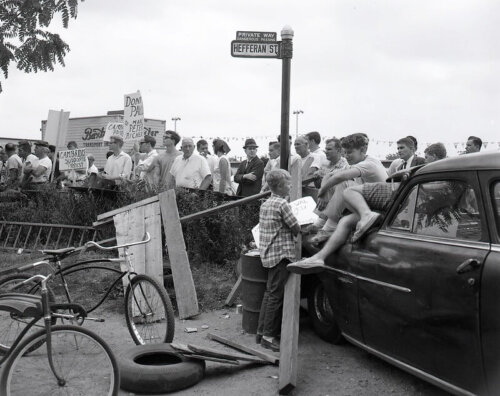 A group of protestors standing behind a fence while a group of young boys sit on the hood of a car next to some bikes.