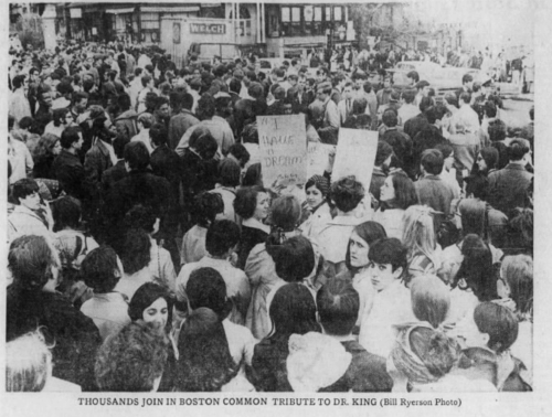 A large crowd in a park with several people holding signs and two women in the lower third looking at the camera.