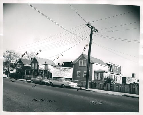 A row of single family homes with a large telephone pole out front. The first house has a sign that says "To hell with urban renewal."