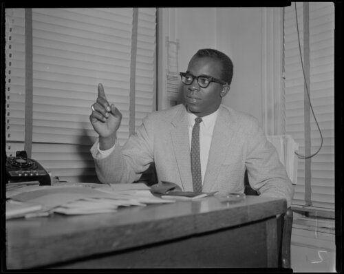 A man sitting at at a desk in front of blinds with his finger pointing up.