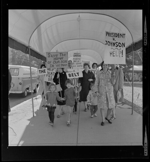 A group of men, women, and children marching with signs underneath an awning.