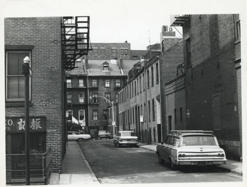 A street in a city with low brick buildings and midcentury cars.
