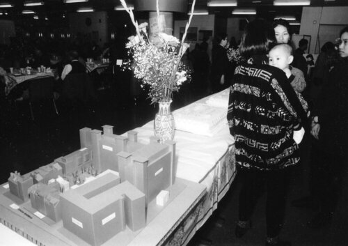 A woman holding a baby faces away from the camera and a table with a vase of flowers and a cardboard architectural model.