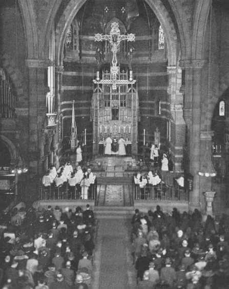 The interior of a church during mass with a choir, parishioners, and priests. There is a large cross hanging above.