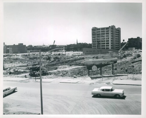 A midcentury car drives by a demolished area with a sign for the new building that is under construction.