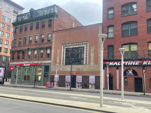 Three brick buildings on a street with the building in the middle with a big central window being the shortest.