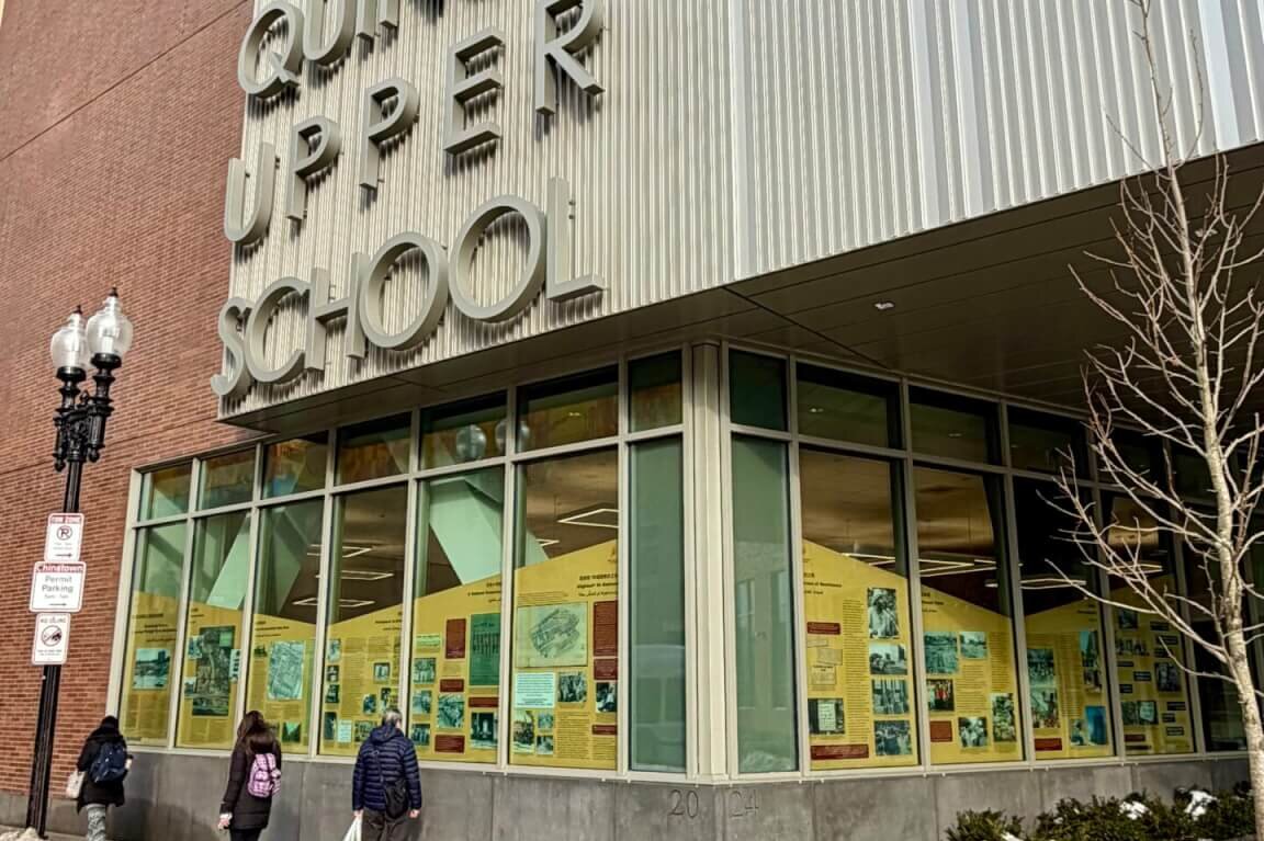 The side of a school building with the windows covered in an exhibit. There are three people walking past next to a pile of snow.