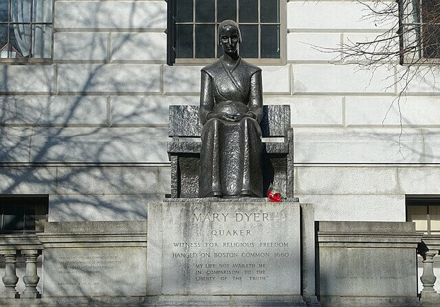 A statue on a pedestal of a woman wearing a head covering and seated on a bench.