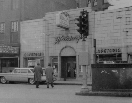 The front of a building with the word Waldorf over the door. Two men walk towards the door.