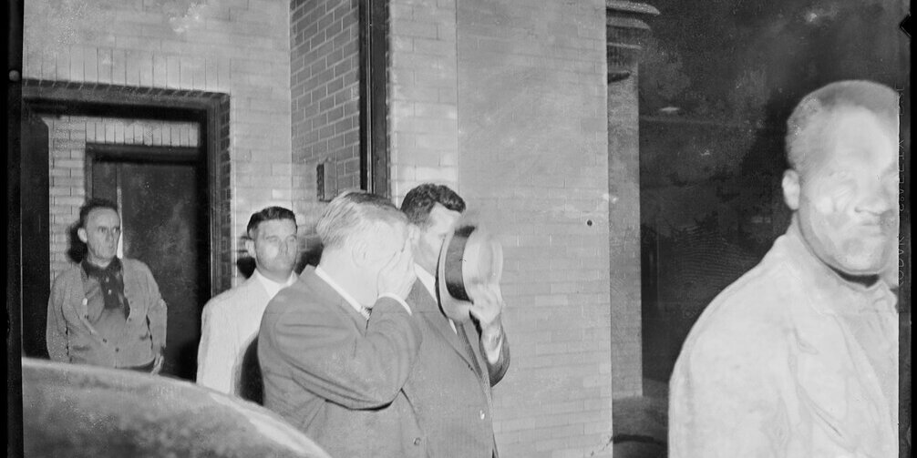 Two men covering their faces with a hat and hand against the camera as they are escorted out of a courthouse.