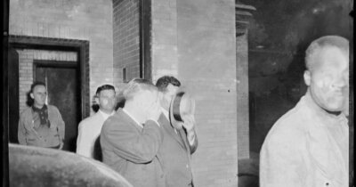 Two men covering their faces with a hat and hand against the camera as they are escorted out of a courthouse.