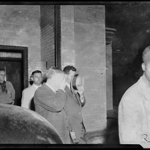 Two men covering their faces with a hat and hand against the camera as they are escorted out of a courthouse.