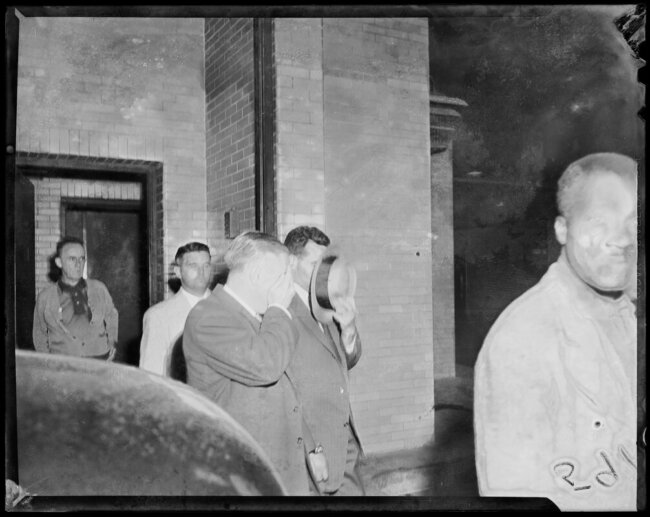 Two men covering their faces with a hat and hand against the camera as they are escorted out of a courthouse.