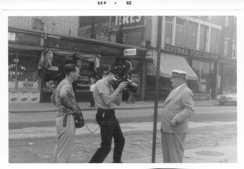 Three men with film equipment standing in front of storefronts.