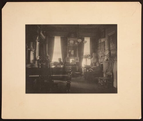 An early twentieth century parlor with a piano in the foreground and art all over the walls. Two women sit in chairs writing and reading.