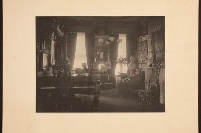 An early twentieth century parlor with a piano in the foreground and art all over the walls. Two women sit in chairs writing and reading.