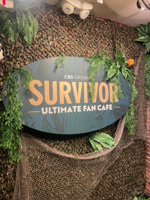 A sign that says "Survivor Ultimate Fan Cafe" with camo netting and fake foliage around the sign.