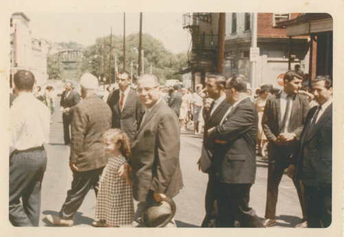 A group of men in their Sunday best walking in procession across the street.