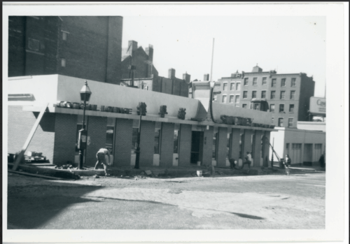 A black and white image of a low building with chinese characters on the side next to brick buildings.