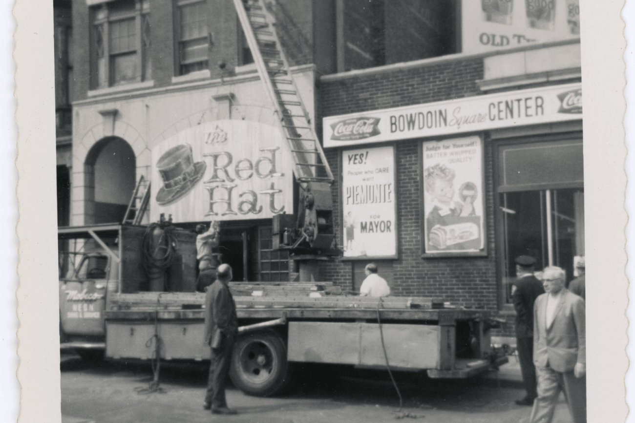 A black and white photo of a truck and crane hanging up a sign with a large hat painted on it with the words "Red Hat."