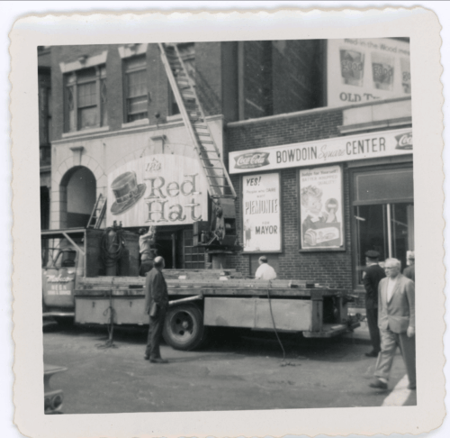 A black and white photo of a truck and crane hanging up a sign with a large hat painted on it with the words "Red Hat."