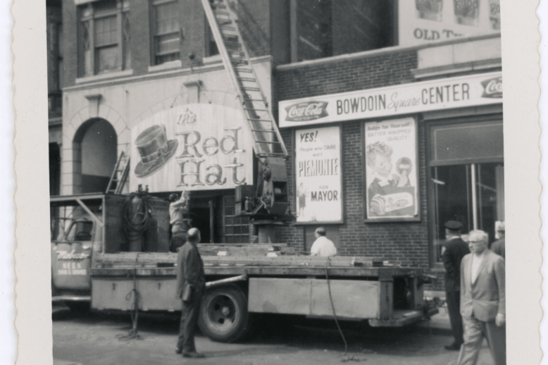 A black and white photo of a truck and crane hanging up a sign with a large hat painted on it with the words "Red Hat."