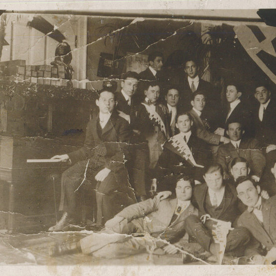 A group of young men posing around a piano. The photo is tattered and over exposed.