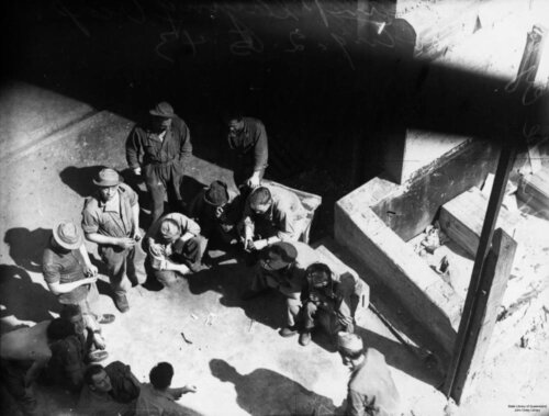 A group of men, some in uniform, stand or squat in a circle playing a game of craps. The photo is taken from above looking down into a construction site.