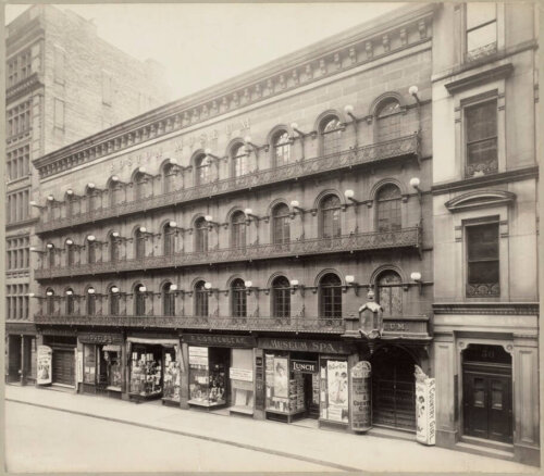 A four story building with elaborate iron balcony on the upper stories. The first floor has shop windows. The words Boston Museum are along the top.