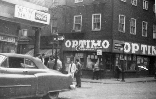 A busy street corner with the left side covered by a 1950s car. The right side of the street has a cigar shop and the right side has a lunch counter.