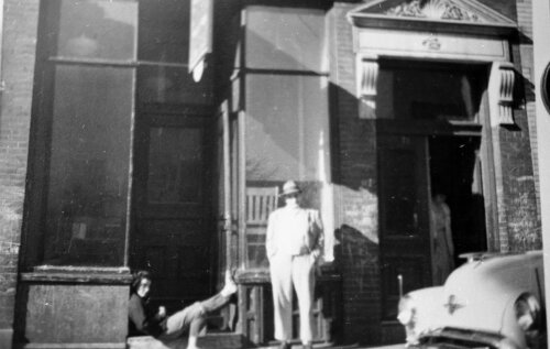 A man standing in front of a shop window while another person lounges on the stoop with their feet propped up on the front step.