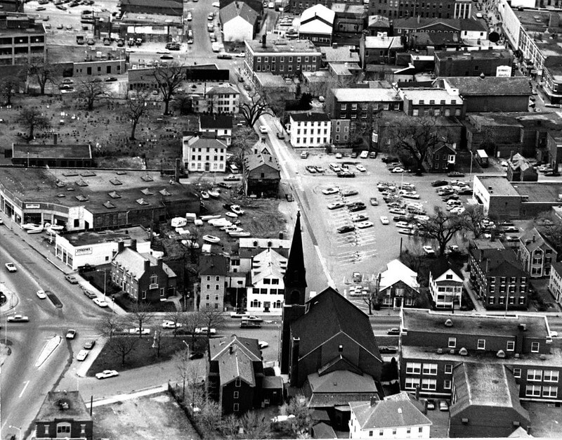 An aerial view of a downtown area. A church can be seen in the bottom third in the center and a parking lot in the middle.