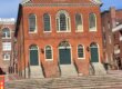 A three story brick building in a federal style with large arched windows and doors facing a paved courtyard.