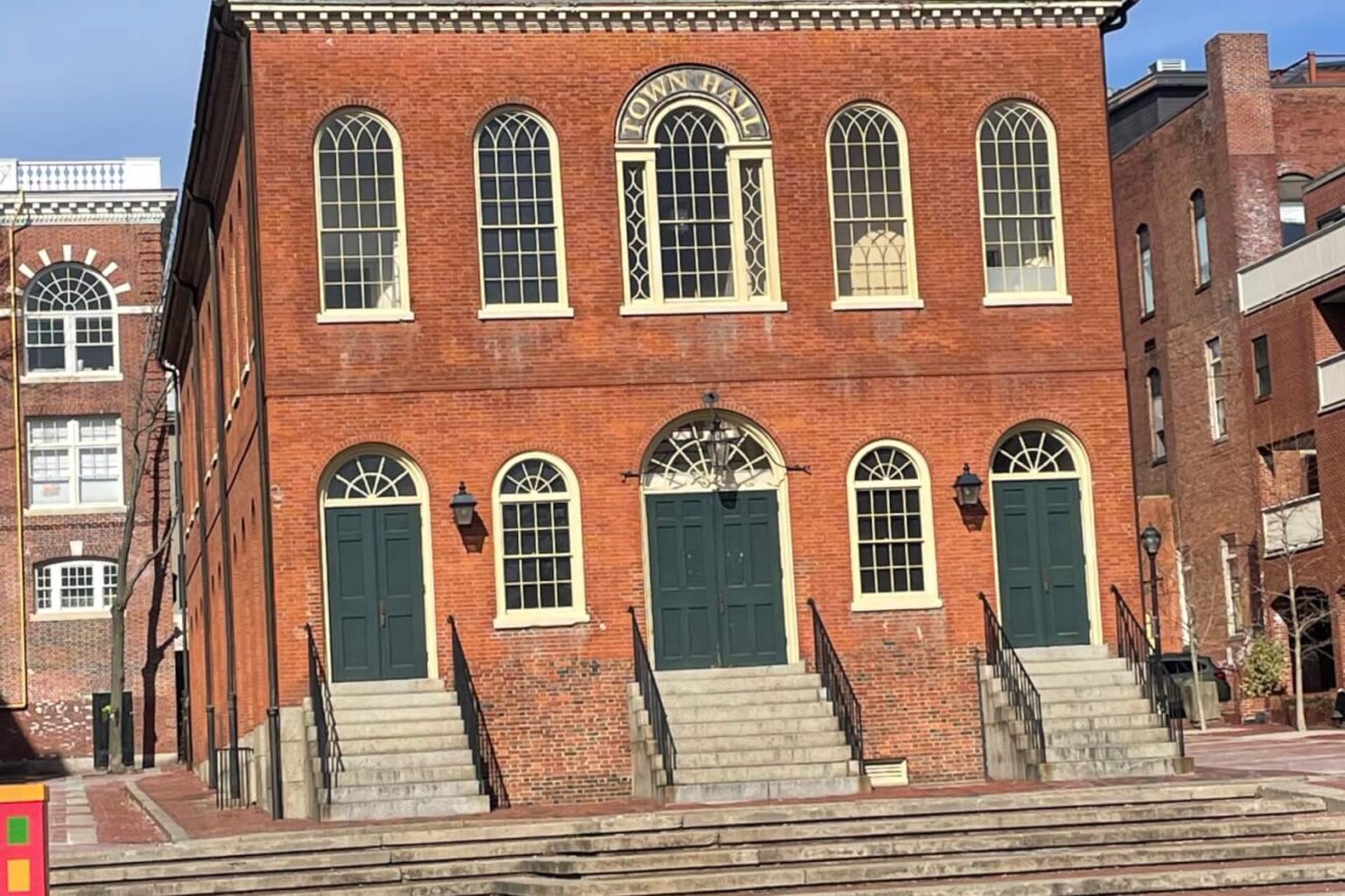 A three story brick building in a federal style with large arched windows and doors facing a paved courtyard.