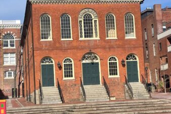 A three story brick building in a federal style with large arched windows and doors facing a paved courtyard.