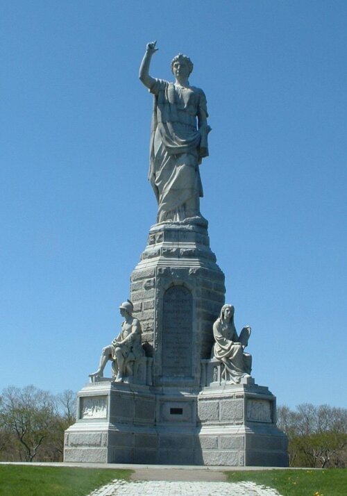 A statue of man in an orator's pose and robes standing on a tall plinth with a soldier and a woman holding a stele sit on a lower level.