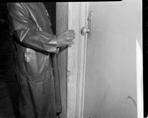 A man wearing a leather coat and glove leans on a closed doorframe.