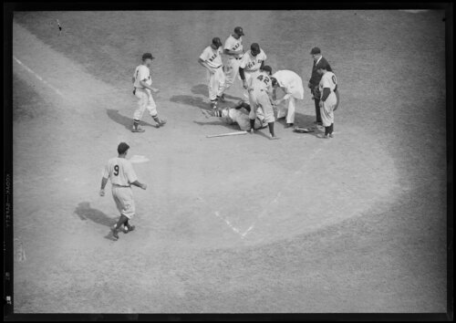 A group of baseball players and an umpire approach a player who has fallen down near home plate.
