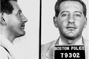 A profile and face on shot of a man with a five o'clock shadow. He has a Boston police prisoner placard around his neck.