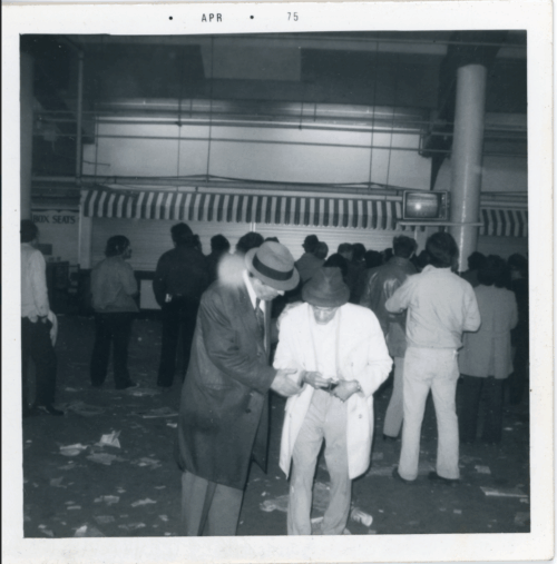 A man in a white coat and hat and a man in a dark coat look at betting sheets at a horse race track. There are many other people around.