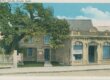 A postcard of a colonial house with a more modern storefront closer to the street. There is a large tree covering most of the hosue