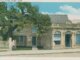 A postcard of a colonial house with a more modern storefront closer to the street. There is a large tree covering most of the hosue